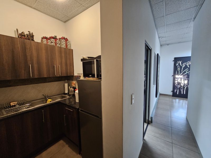 A compact office kitchenette featuring dark wood cabinetry, granite countertops, and a stainless steel sink, adjacent to a tiled hallway with decorative security door.