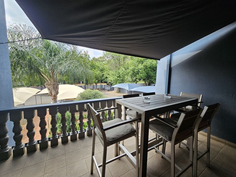 A covered balcony featuring a balustrade railing and a retractable shade canopy, overlooking a landscaped outdoor area with parking shade structures.