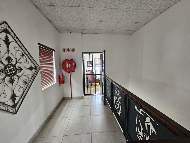A bright office hallway featuring tiled flooring, a suspended ceiling, a security gate, and wall-mounted fire safety equipment.