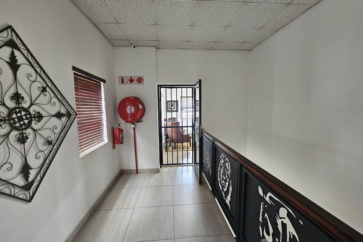 A bright office hallway featuring tiled flooring, a suspended ceiling, a security gate, and wall-mounted fire safety equipment.