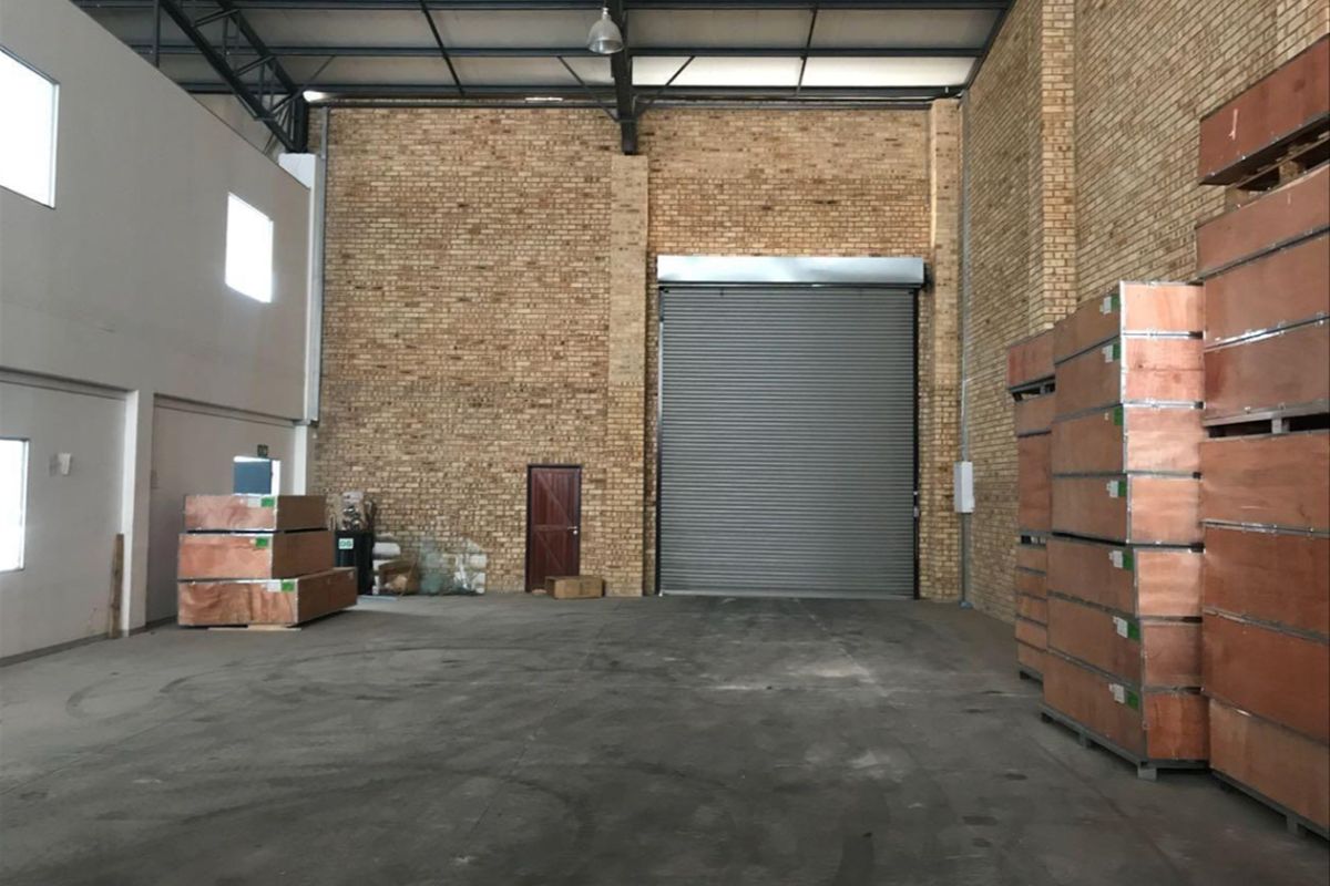Spacious industrial warehouse unit featuring high exposed brick walls, concrete flooring, steel roof trusses, and a large roller shutter door for easy access.