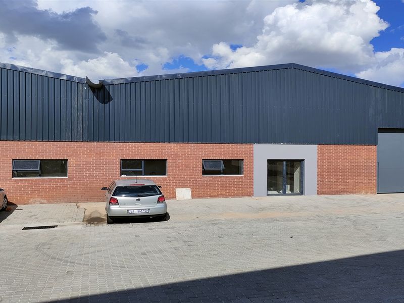 Industrial warehouse exterior featuring a brick facade, corrugated metal roofing, paved driveway, and large industrial roller shutter door access.
