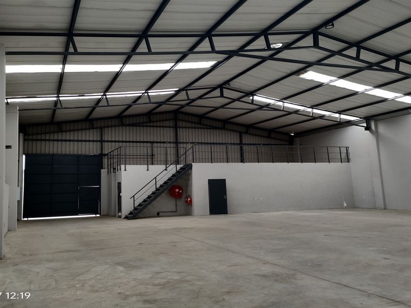 Spacious industrial warehouse featuring a high-clearance steel truss roof, concrete flooring, a mezzanine level with staircase access, and a large roller shutter door.