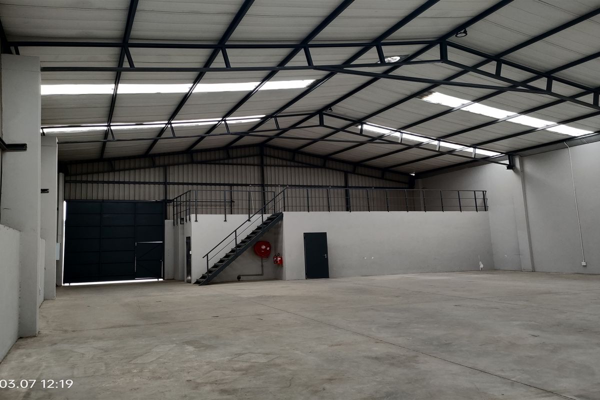 Spacious industrial warehouse featuring a high-clearance steel truss roof, concrete flooring, a mezzanine level with staircase access, and a large roller shutter door.