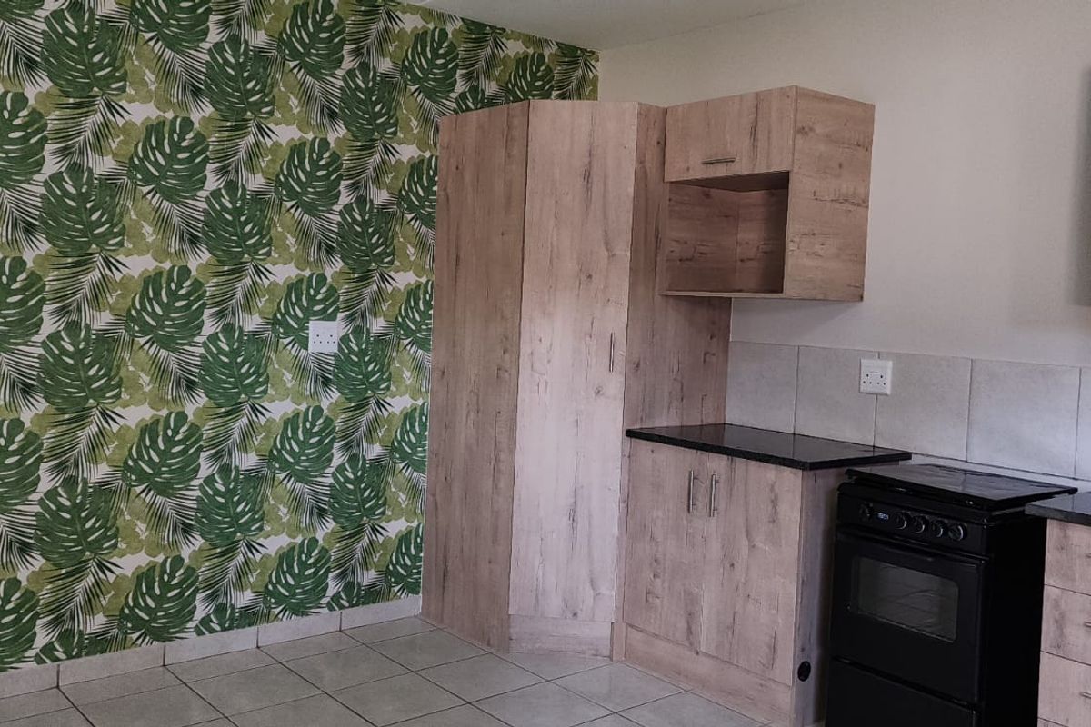 A compact office kitchenette featuring light wood-grain cabinetry, dark countertops, tiled flooring, and a decorative botanical wallpaper feature wall.