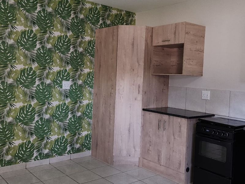 A compact office kitchenette featuring light wood-grain cabinetry, dark countertops, tiled flooring, and a decorative botanical wallpaper feature wall.