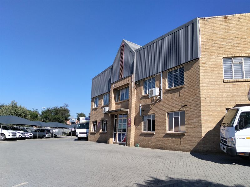 Front elevation of a multi-story industrial building featuring face brick construction, metal roof cladding, and an adjacent paved parking area with covered bays.