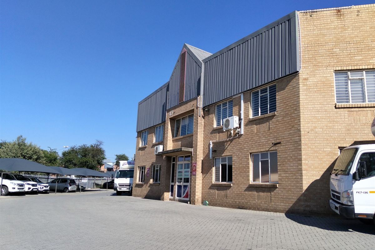 Front elevation of a multi-story industrial building featuring face brick construction, metal roof cladding, and an adjacent paved parking area with covered bays.