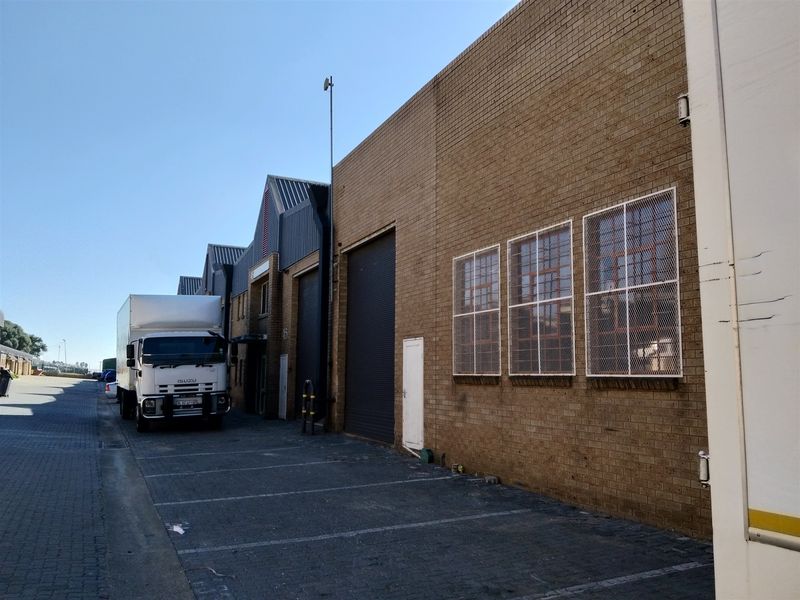 Industrial warehouse exterior featuring a brick facade, roller shutter door, and security-screened windows with adjacent paved parking area.