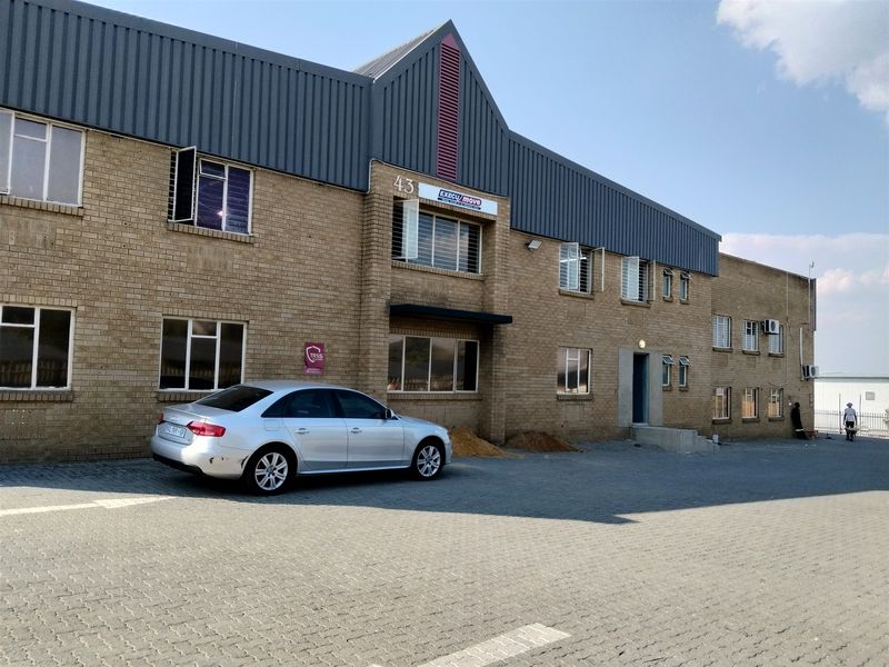 Front elevation of a multi-story industrial building featuring face brick construction, corrugated metal roofing, and an expansive paved parking area.