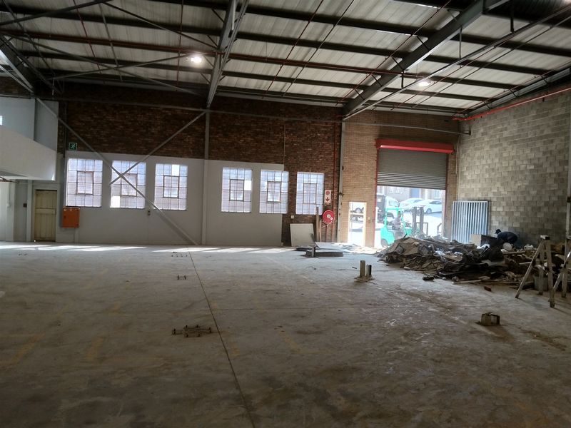 Spacious industrial warehouse unit featuring high ceilings with exposed steel trusses, concrete flooring, and a large roller shutter door for vehicle access.