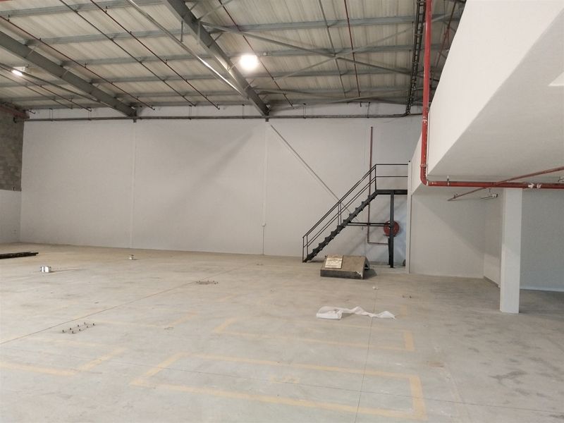 Spacious industrial warehouse interior featuring high ceilings with exposed steel trusses, concrete flooring, fire sprinkler system, and a metal staircase leading to a mezzanine level.