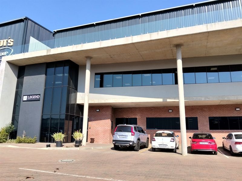 Modern industrial building facade featuring a combination of brickwork, glass curtain walling, and a covered parking area supported by concrete columns.