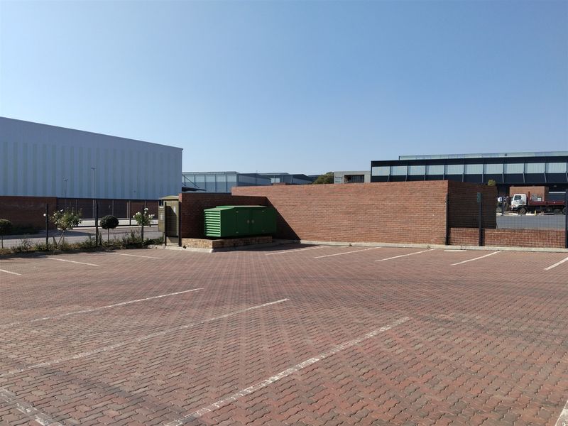 A paved industrial parking area featuring a brick boundary wall, security fencing, and an enclosed electrical transformer unit.