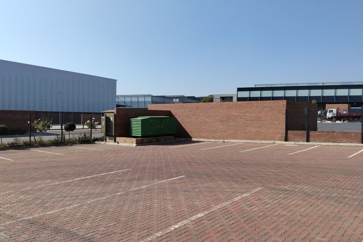 A paved industrial parking area featuring a brick boundary wall, security fencing, and an enclosed electrical transformer unit.