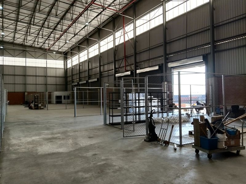 Spacious industrial warehouse featuring high ceilings with exposed steel trusses, concrete flooring, and secure chain-link partitioned storage areas.