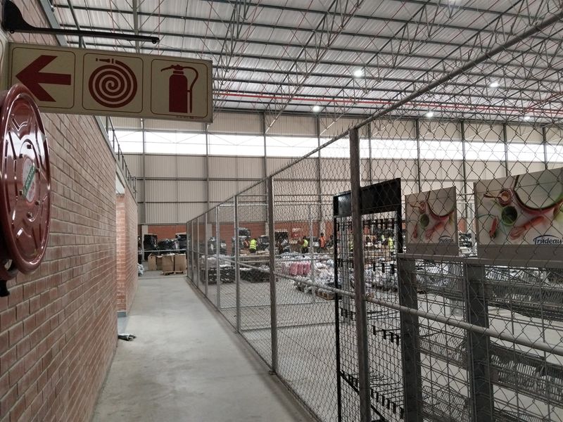 Industrial warehouse interior featuring high ceilings with exposed steel trusses, concrete flooring, fire safety equipment, and partitioned storage areas with chain-link fencing.