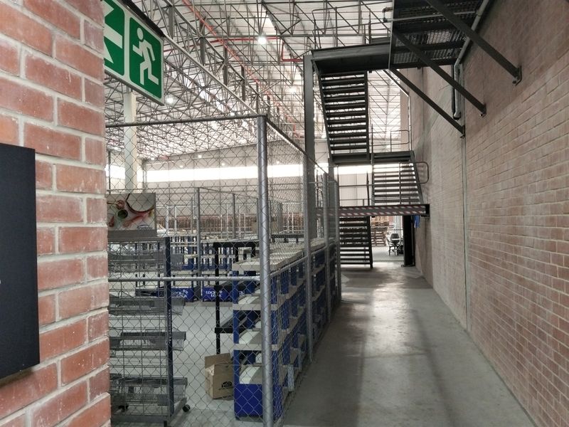 Industrial warehouse space featuring exposed brick walls, high-clearance steel roof trusses, concrete flooring, and a metal staircase with security fencing.