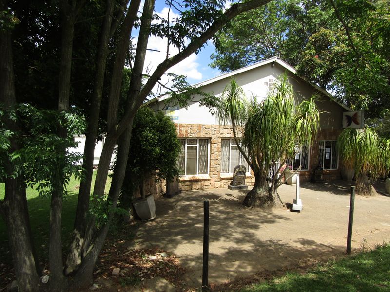 Front exterior view of an office building featuring stone cladding, security window bars, and a paved entrance area surrounded by mature trees.