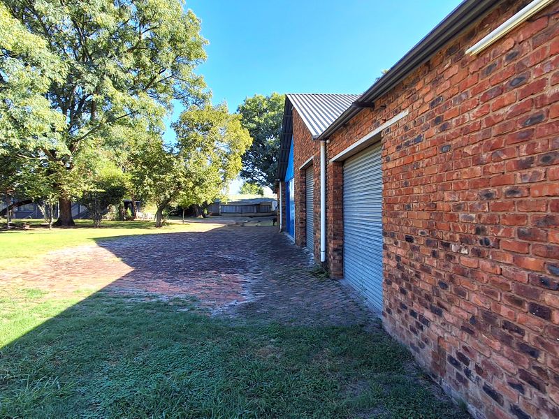 Rear view of the farm property featuring a brick structure with multiple roller shutter doors and an expansive paved area leading to the garden.