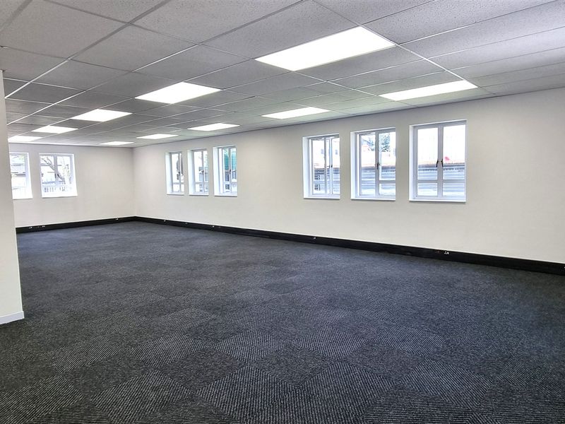 A spacious, open-plan office area featuring neutral walls, carpeted flooring, and ample natural light from multiple windows, suitable for a professional workspace.