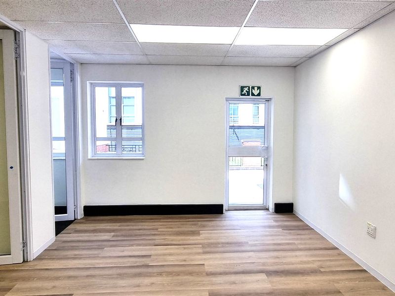 A bright, empty office space featuring wood-look flooring, a suspended ceiling with recessed lighting, and a glass partition door.