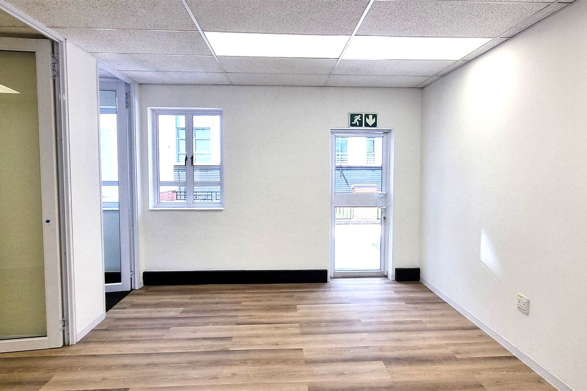 A bright, empty office space featuring wood-look flooring, a suspended ceiling with recessed lighting, and a glass partition door.