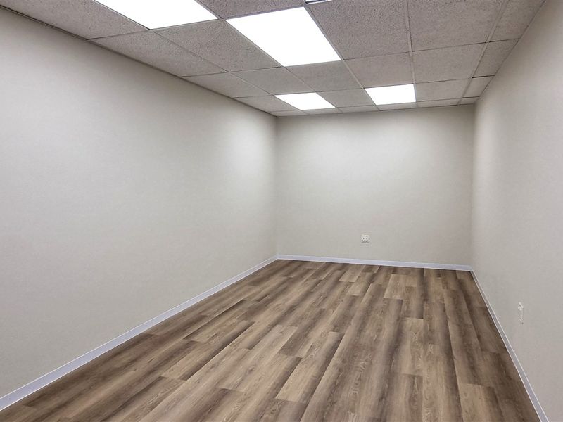 A clean, vacant office space featuring modern laminate wood flooring, neutral walls, and a drop ceiling with integrated LED panel lighting.