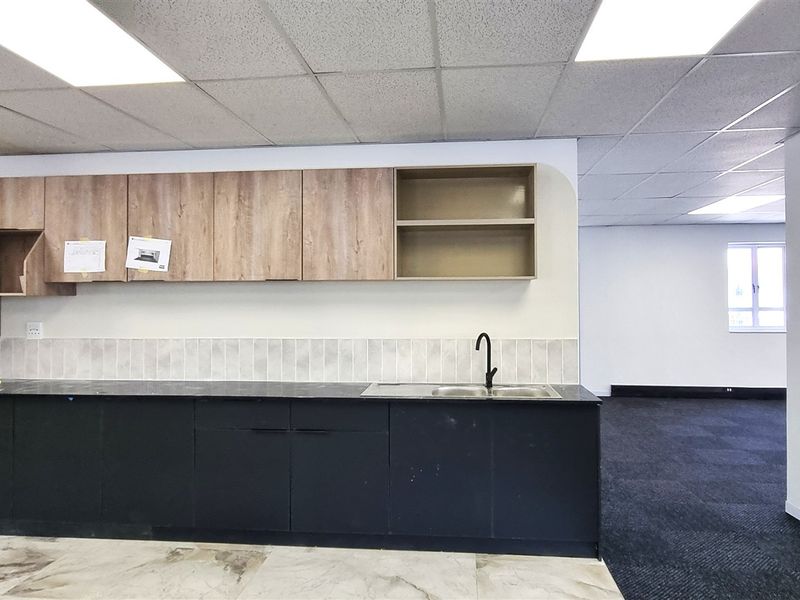 A clean, modern office kitchenette featuring dark base cabinets, wood-tone upper cabinetry, a stainless steel sink, and a mix of tiled and carpeted flooring.