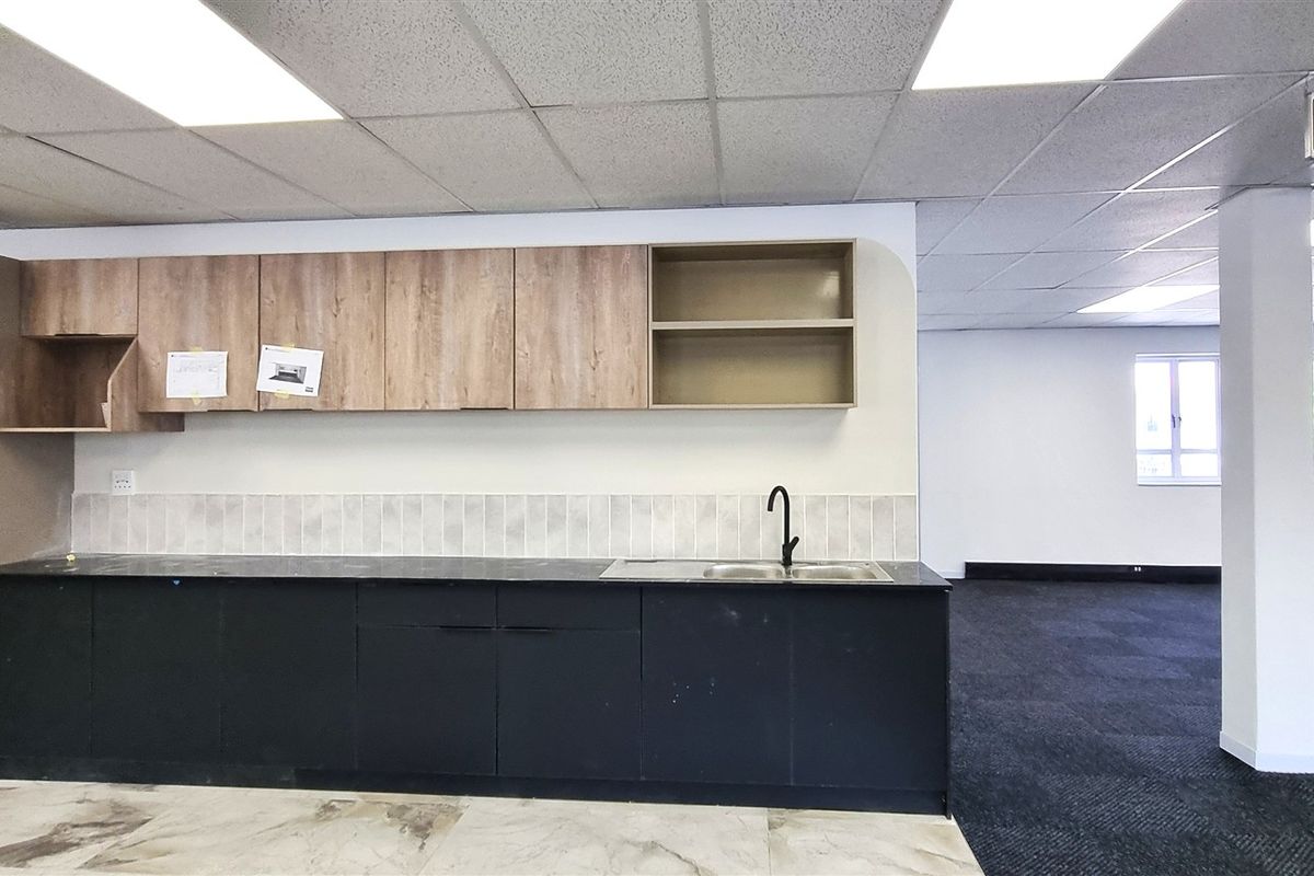 A clean, modern office kitchenette featuring dark base cabinets, wood-tone upper cabinetry, a stainless steel sink, and a mix of tiled and carpeted flooring.