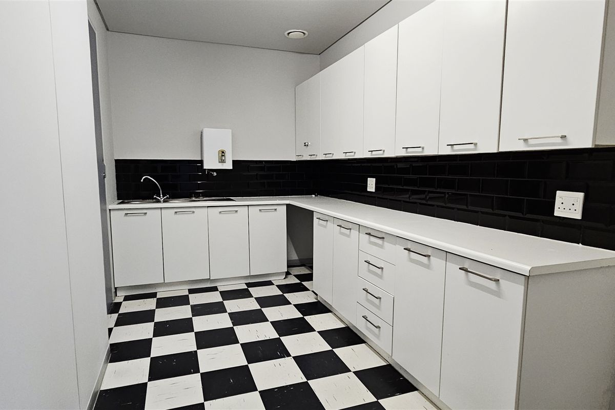 A clean, functional office kitchenette featuring white cabinetry, a black subway tile backsplash, and distinctive checkerboard flooring.