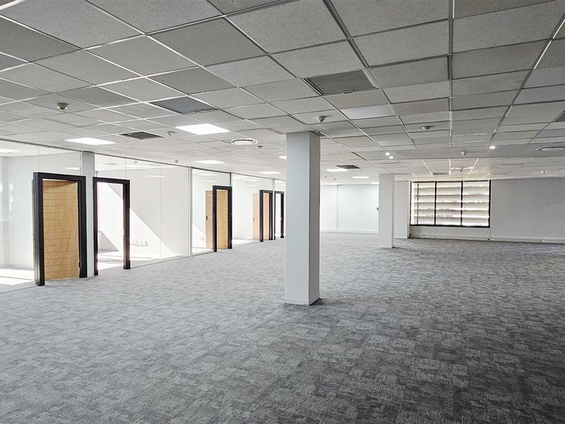 Spacious open-plan office area featuring glass-partitioned private offices, suspended ceilings with recessed lighting, and neutral carpet flooring.