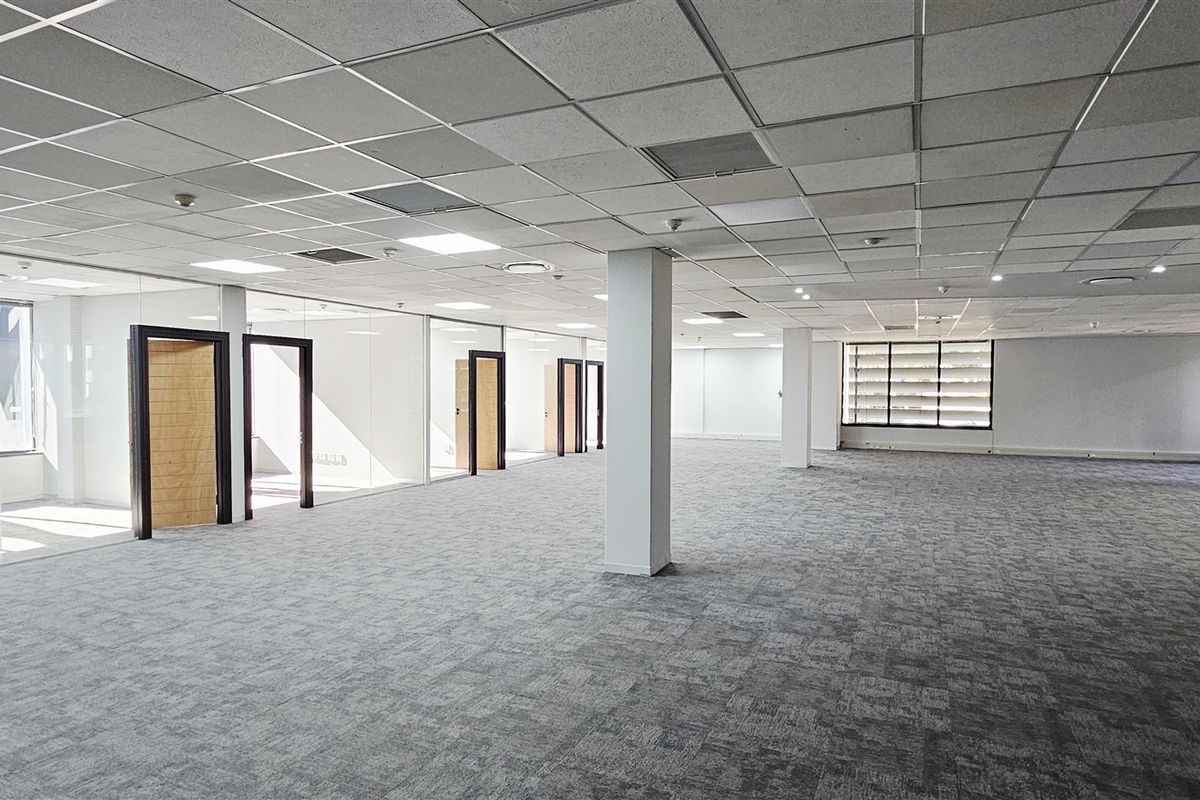 Spacious open-plan office area featuring glass-partitioned private offices, suspended ceilings with recessed lighting, and neutral carpet flooring.