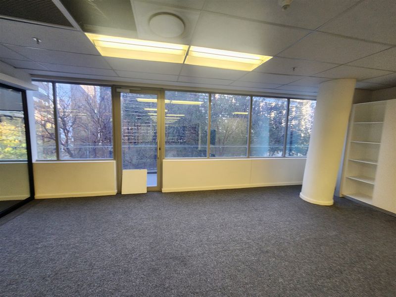 A vacant office space featuring large windows, built-in shelving, a structural column, and carpeted flooring, offering a bright and professional work environment.