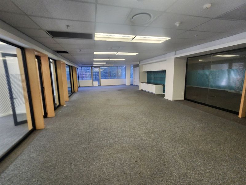 Spacious open-plan office area featuring glass partitions, a built-in kitchenette, suspended ceilings with recessed lighting, and carpeted flooring throughout.