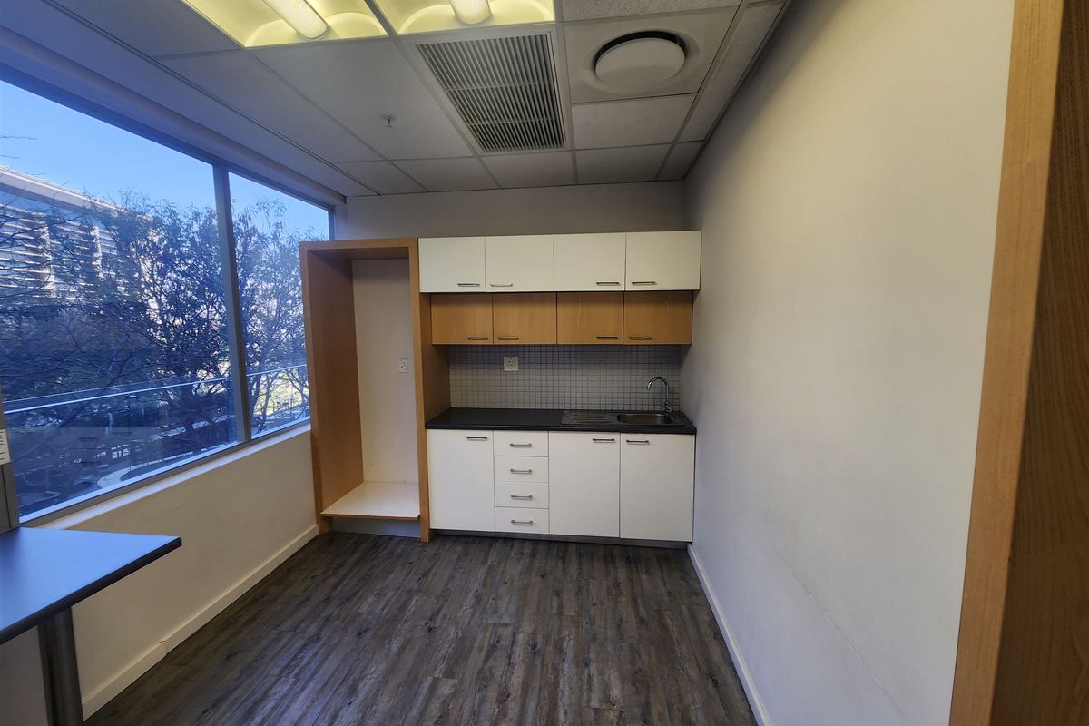 A clean, functional office kitchenette featuring built-in cabinetry, a stainless steel sink, and large windows providing natural light.