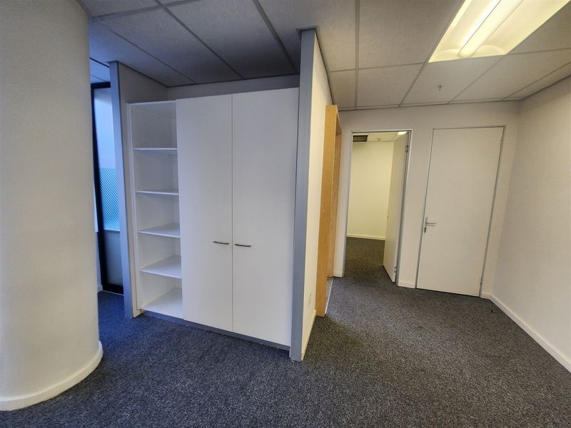 A clean, empty office space featuring built-in storage cabinetry, grey carpet flooring, and a suspended ceiling with integrated lighting.