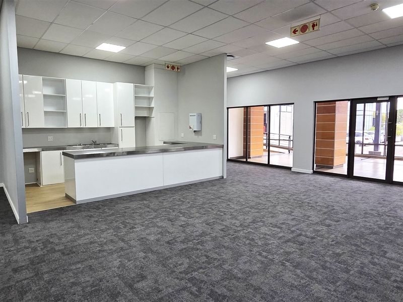 Spacious open-plan office area featuring a modern kitchenette with white cabinetry, stainless steel counters, carpeted flooring, and large glass doors providing natural light.
