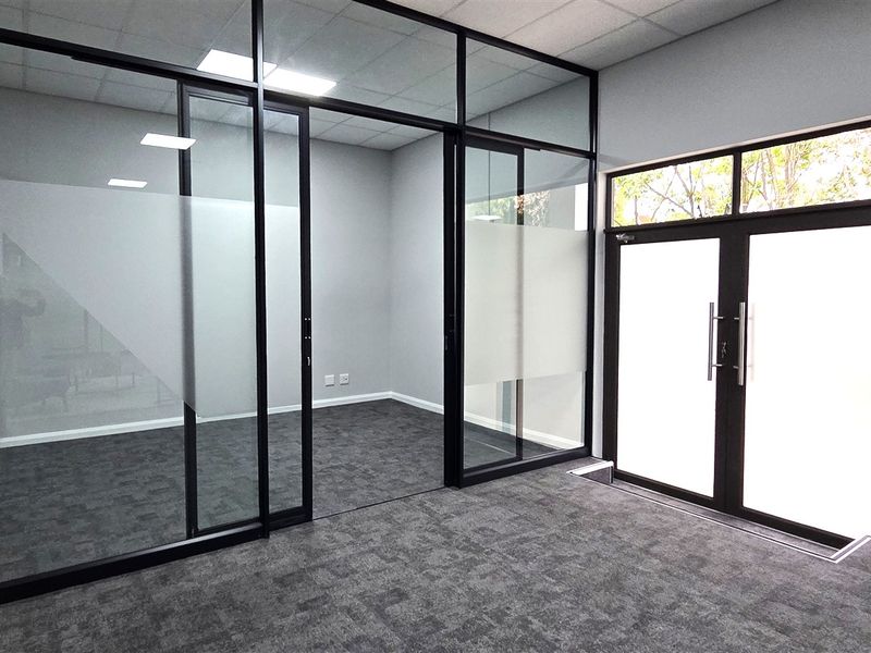 Modern office space featuring glass partition walls with black aluminum frames, carpeted flooring, and a clean, professional aesthetic.