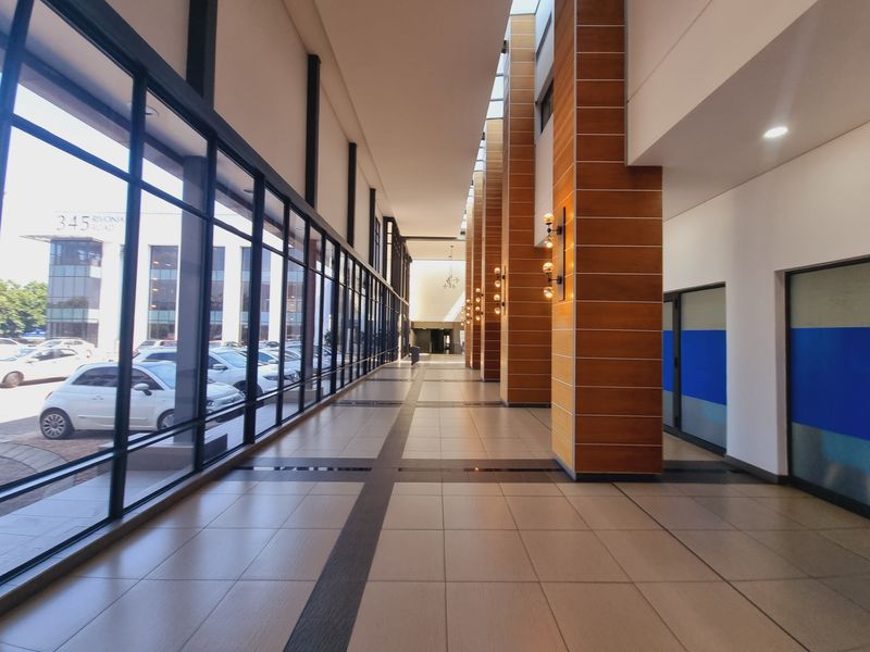 A spacious, modern office building hallway featuring large glass windows, tiled floors, and wood-clad structural columns with integrated lighting.