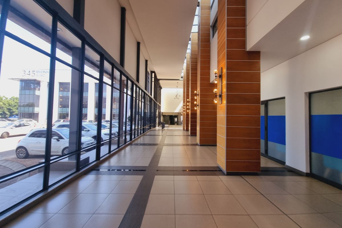 A spacious, modern office building hallway featuring large glass windows, tiled floors, and wood-clad structural columns with integrated lighting.