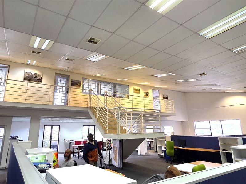 Spacious open-plan office area featuring a mezzanine level, metal staircase, suspended ceilings with recessed lighting, and built-in workstation partitions.