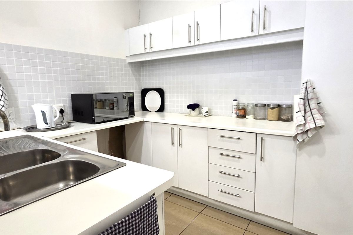 A functional office kitchen featuring white cabinetry, laminate countertops, a double bowl stainless steel sink, and a tiled backsplash.