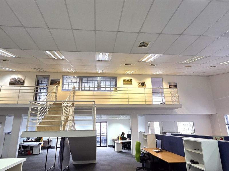 Spacious open-plan office area featuring a mezzanine level, metal staircase, suspended ceilings with recessed lighting, and integrated workstation partitions.