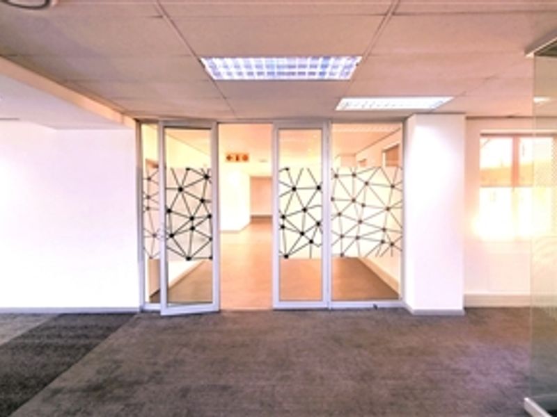 Spacious open-plan office area featuring modern glass partitions, suspended ceilings with recessed lighting, and neutral carpet flooring.