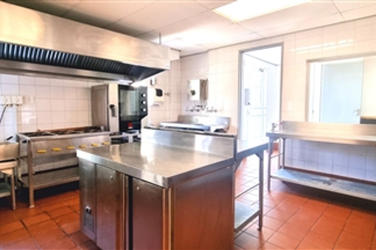 A functional commercial kitchen space featuring stainless steel prep surfaces, a large extractor hood, gas range, and durable tiled finishes throughout.