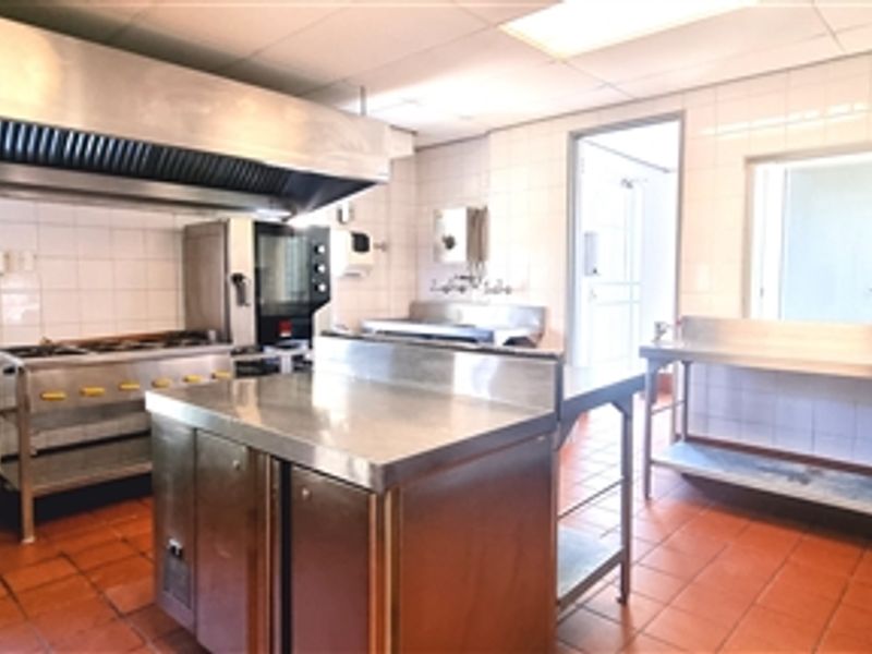 A functional commercial kitchen space featuring stainless steel prep surfaces, a large extractor hood, gas range, and durable tiled finishes throughout.