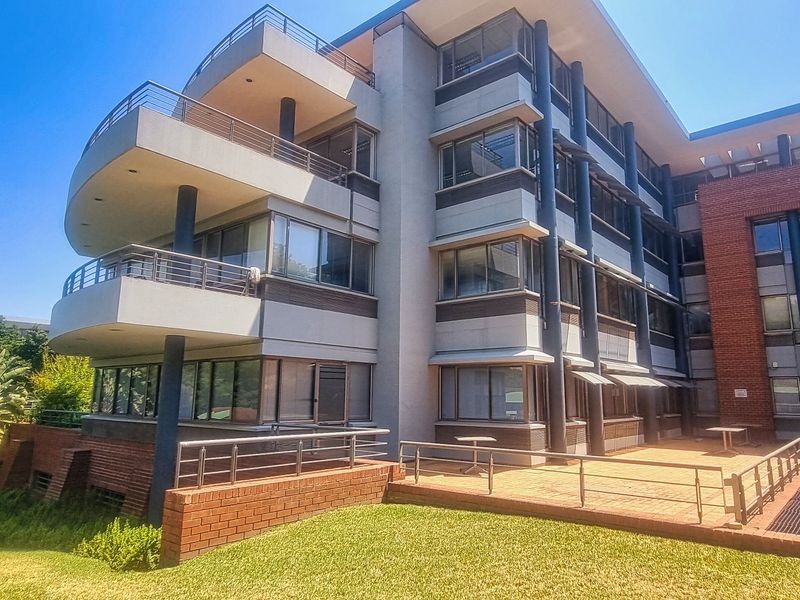 Rear view of a multi-story office building featuring tiered balconies, brick and concrete facade, and a paved patio area overlooking a landscaped garden.