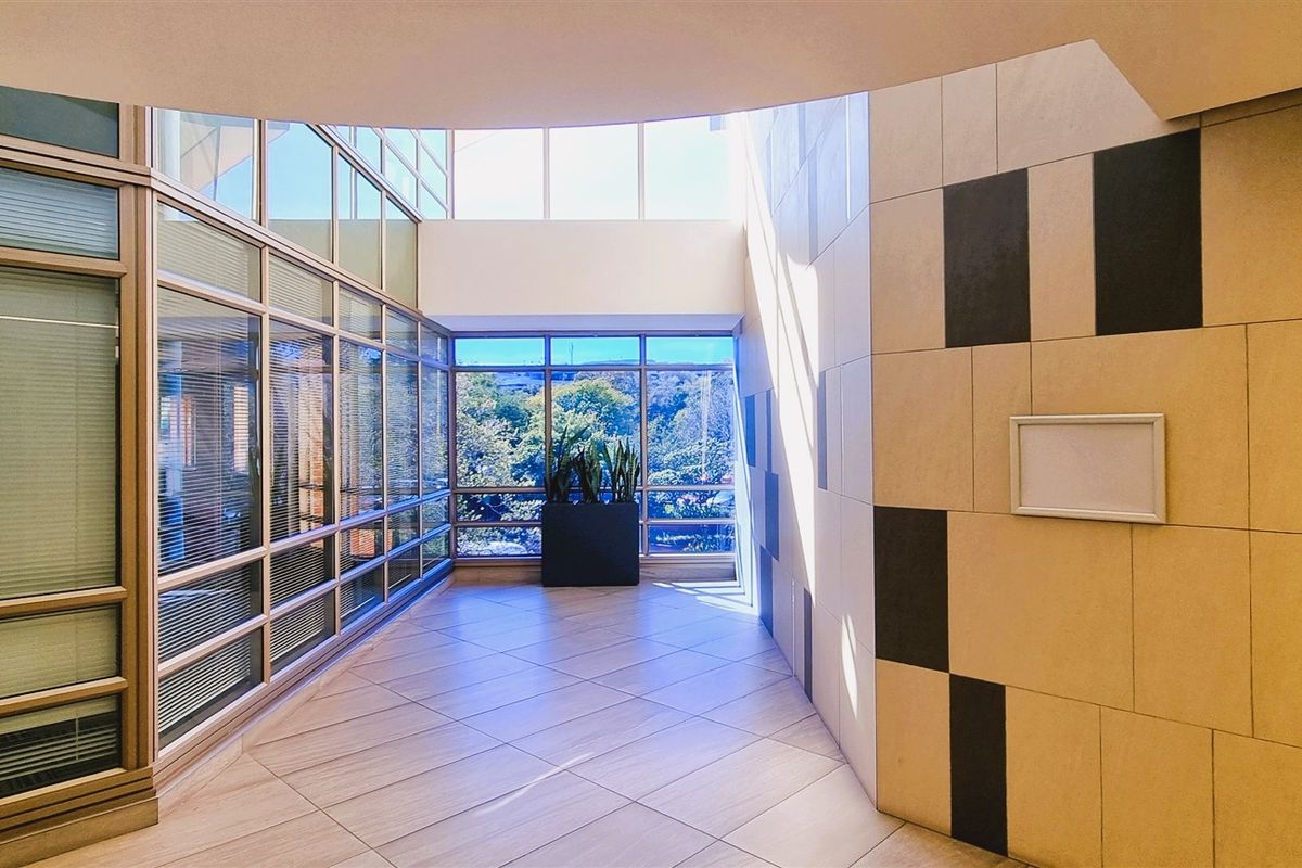 Modern office lobby or circulation area featuring extensive glass curtain walls, natural light from a skylight, and contemporary tiled wall finishes.