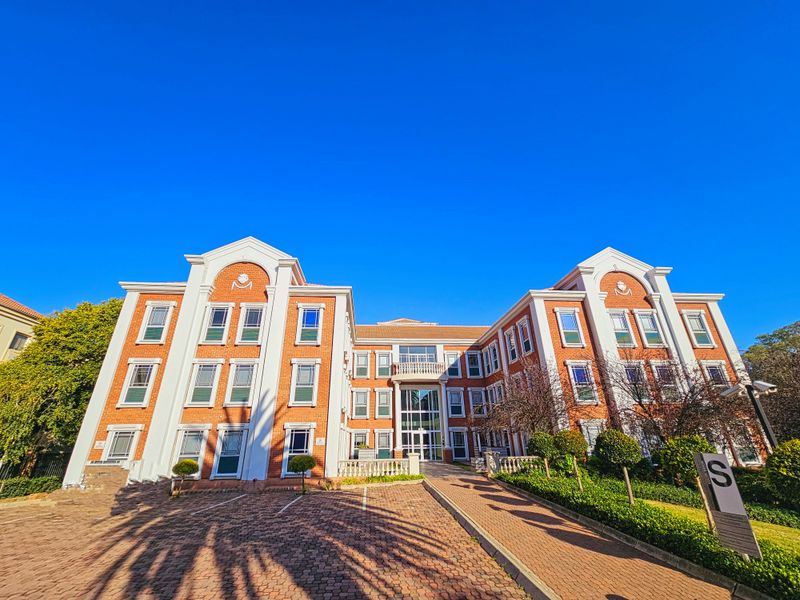 A well-maintained multi-story office building featuring a classic brick facade, paved driveway, and landscaped grounds under a clear blue sky.